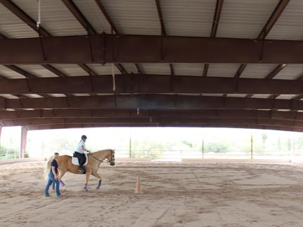 Student riding in covered arena, instructor walking beside